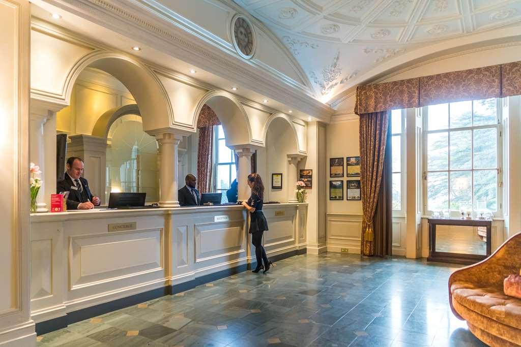 Macdonald Bath Spa Hotel Interior foto The photo shows the interior of a hotel reception area. There is a stylish front desk with several staff members attending to guests. A woman is interacting with one of the receptionists. The design features elegant architectural details, including a