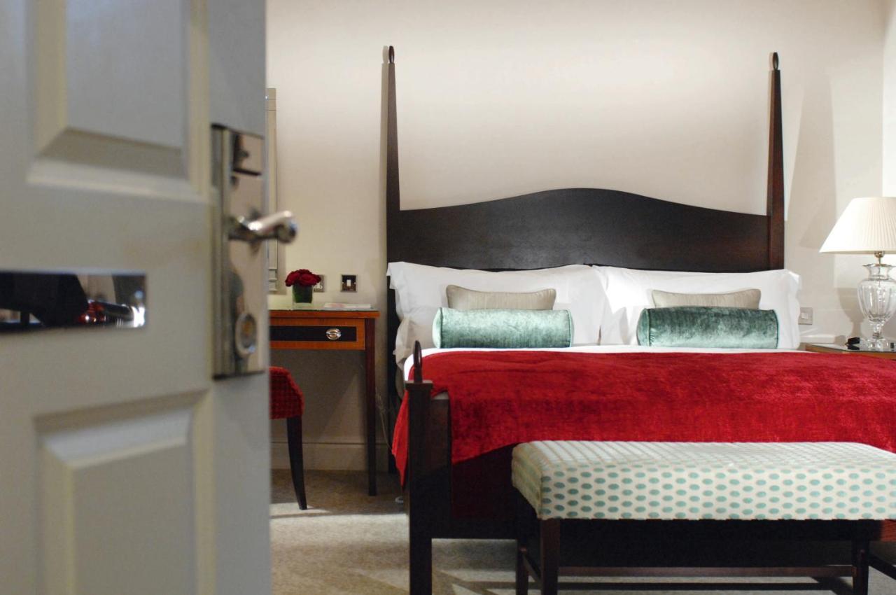 Macdonald Bath Spa Hotel Zimmer foto The photo shows a well-decorated hotel room. In the foreground, there is a partially opened door leading into the room. The focal point of the room is a large bed with a dark wooden headboard and stylish red and white bedding, accented with green pil