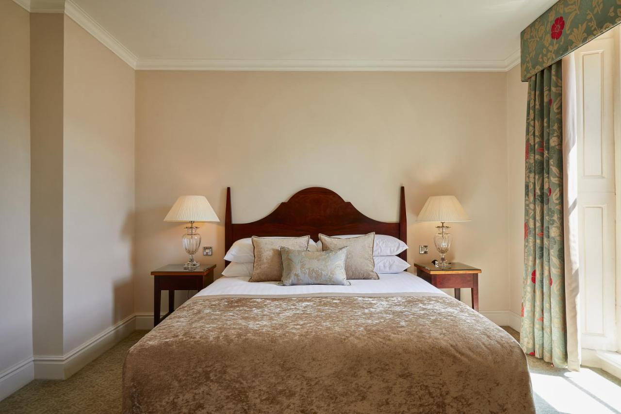 Macdonald Bath Spa Hotel Exterior foto The photo shows a cozy hotel bedroom. It features a large bed with a plush, light-colored bedspread and several pillows. On either side of the bed, there are wooden nightstands with lamps providing soft lighting. The walls are painted a neutral color