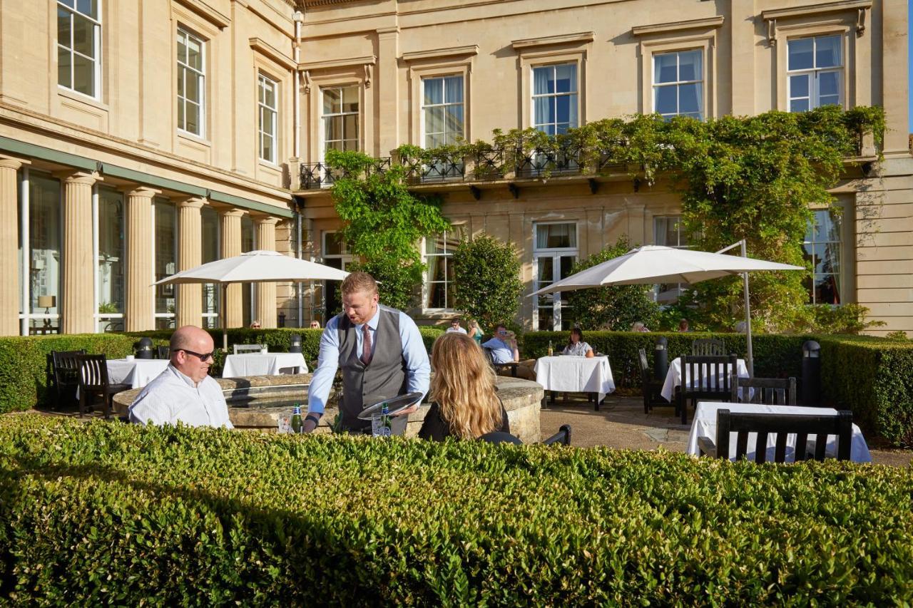 Macdonald Bath Spa Hotel Exterior foto The photo depicts an outdoor dining area of a restaurant. In the foreground, there are neatly trimmed hedges surrounding the dining space. Two patrons are seated at a table; one is a man wearing sunglasses and the other is a woman facing him. A waite