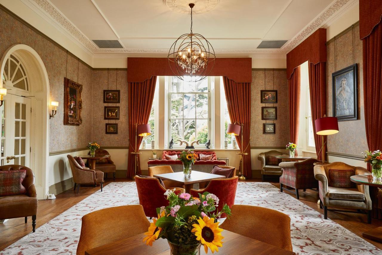 Macdonald Bath Spa Hotel Exterior foto The photo shows a well-decorated living room or lounge area. The space features elegant furnishings, including chairs and a round table at the center. Large windows allow natural light to fill the room, draped with rich red curtains. The walls are ad