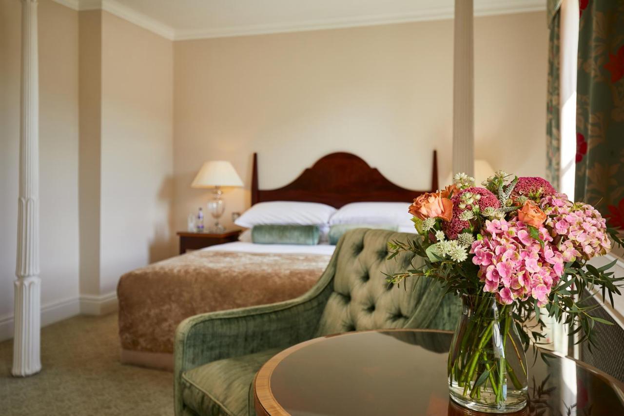 Macdonald Bath Spa Hotel Exterior foto The photo shows a cozy bedroom featuring a large bed with a wooden headboard, topped with a fluffy blanket. To one side, there is a small bedside lamp providing soft lighting. In the foreground, there is an elegant green chair with a tufted design an