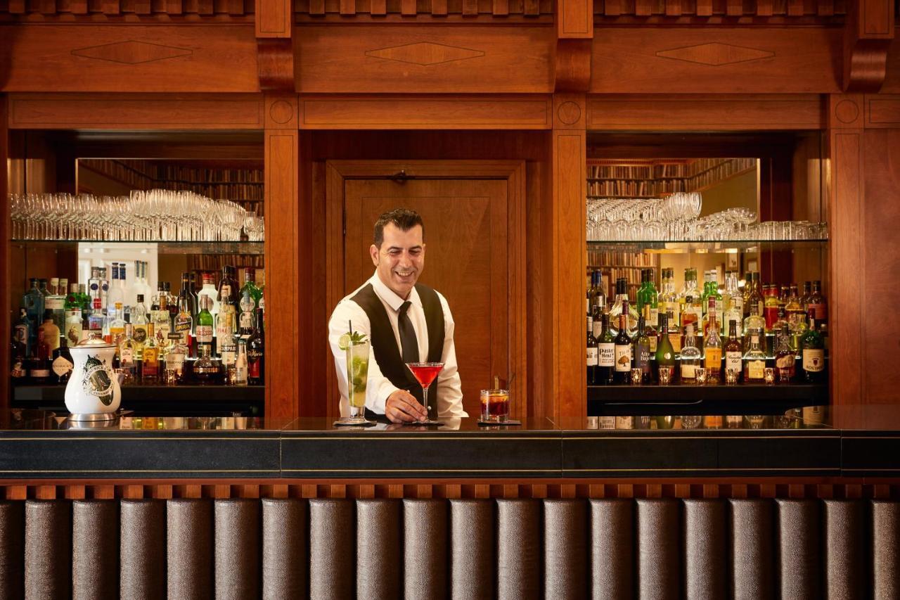 Macdonald Bath Spa Hotel Exterior foto The photo depicts a bartender standing behind a polished bar. He is wearing a formal outfit with a vest and a bow tie, and he appears to be smiling as he presents a cocktail. Behind him is a well-stocked bar shelf filled with various bottles of liquo