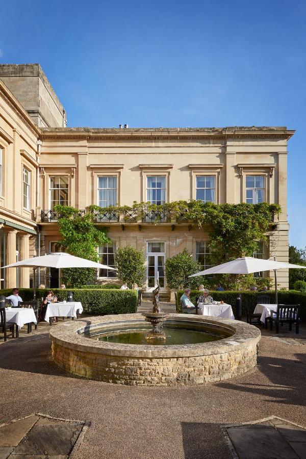 Macdonald Bath Spa Hotel Exterior foto The photo depicts a charming outdoor dining area of a classic-looking building with elegant architecture. The structure features large windows and is adorned with greenery, including climbing plants. In the foreground, there is a circular fountain, a