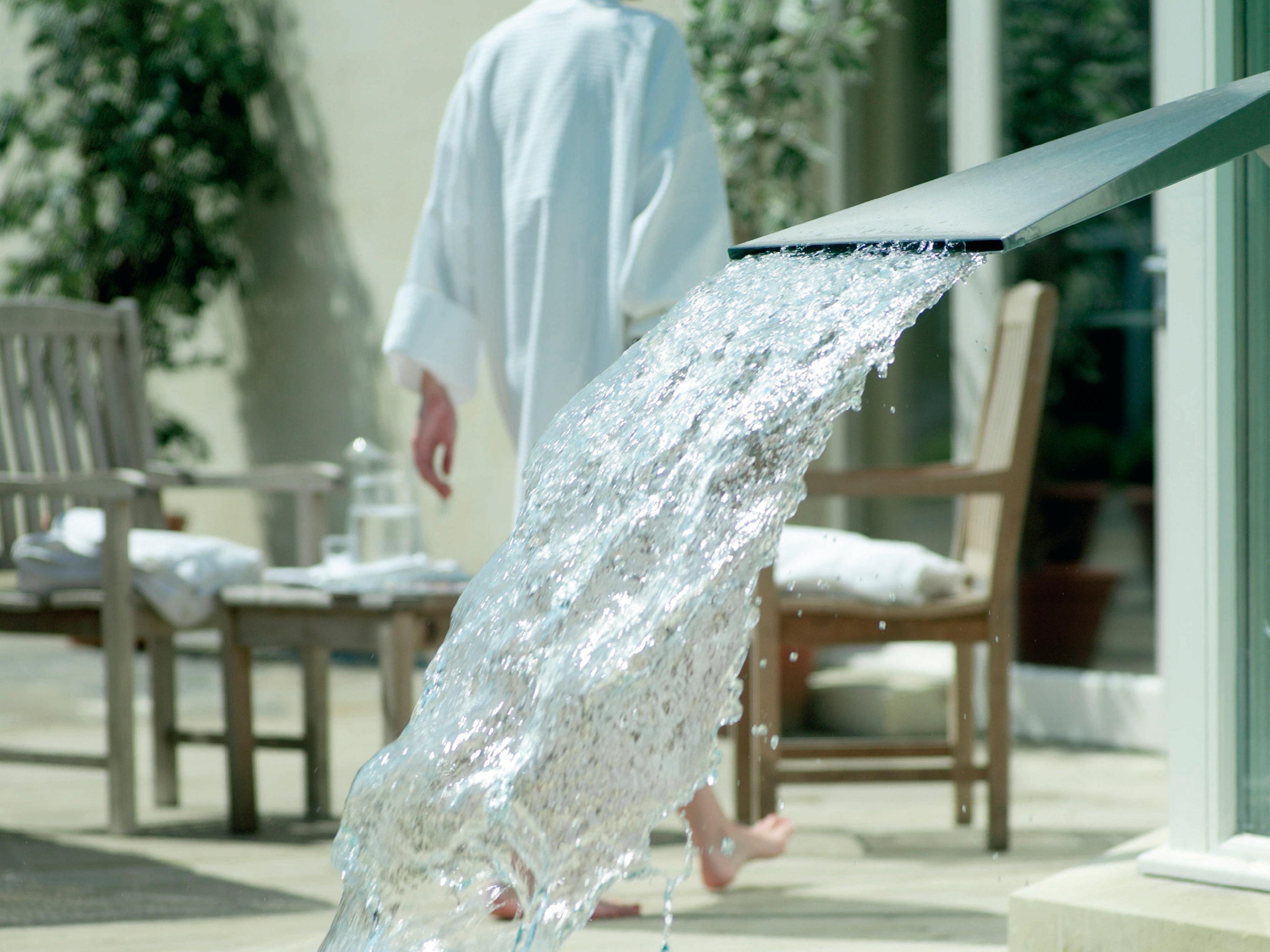 Macdonald Bath Spa Hotel Exterior foto The photo shows a flowing water feature, with water elegantly cascading from a sleek spout. In the background, there is a person wearing a white bathrobe, walking away. Surrounding the area, there are wooden chairs and what appears to be a serene out