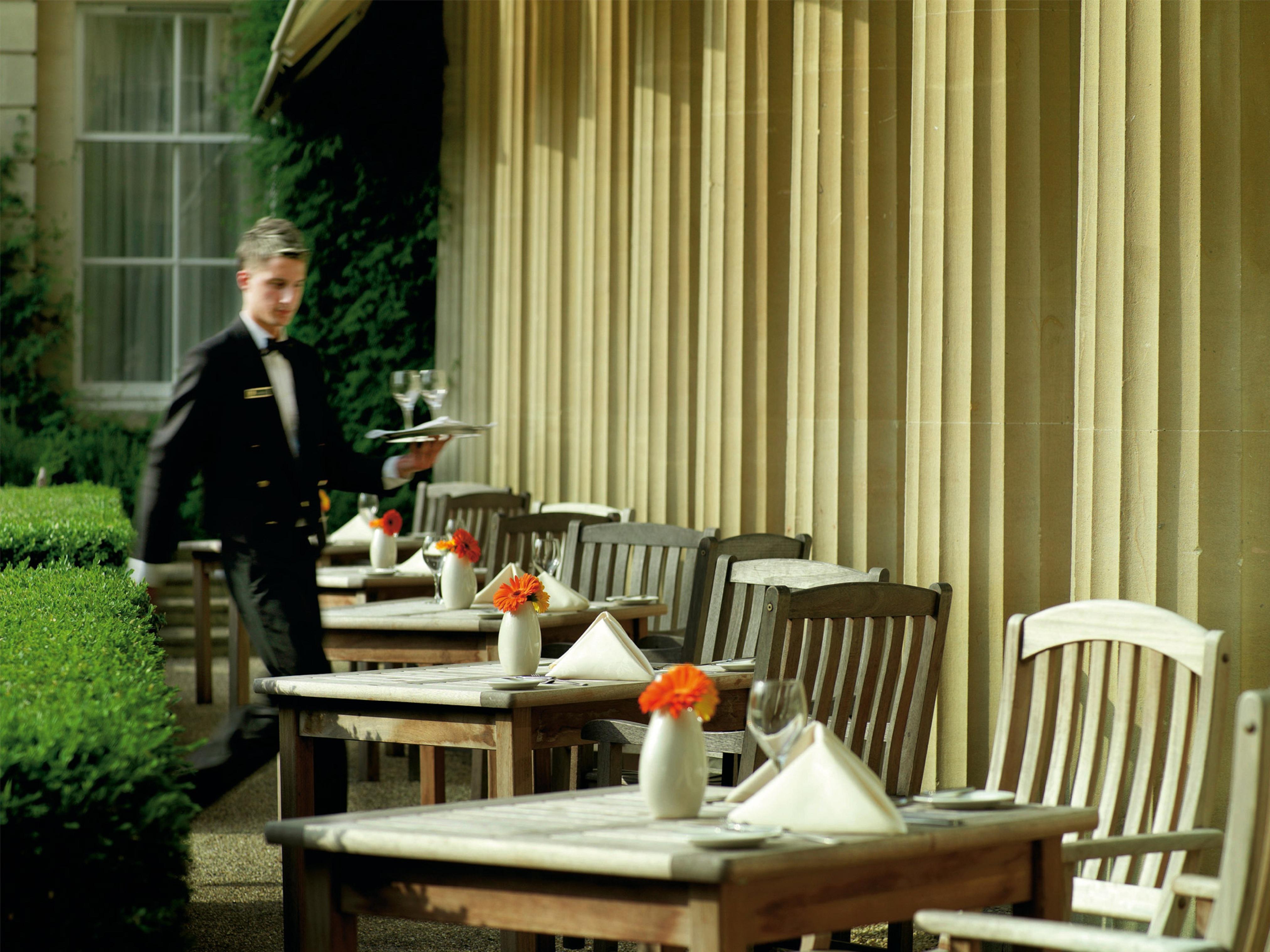 Macdonald Bath Spa Hotel Exterior foto The photo shows an outdoor dining area, featuring several wooden tables and chairs arranged neatly. Each table is set with a white tablecloth and small vases containing flowers, adding a vibrant touch. A waiter in a formal black suit is seen moving t