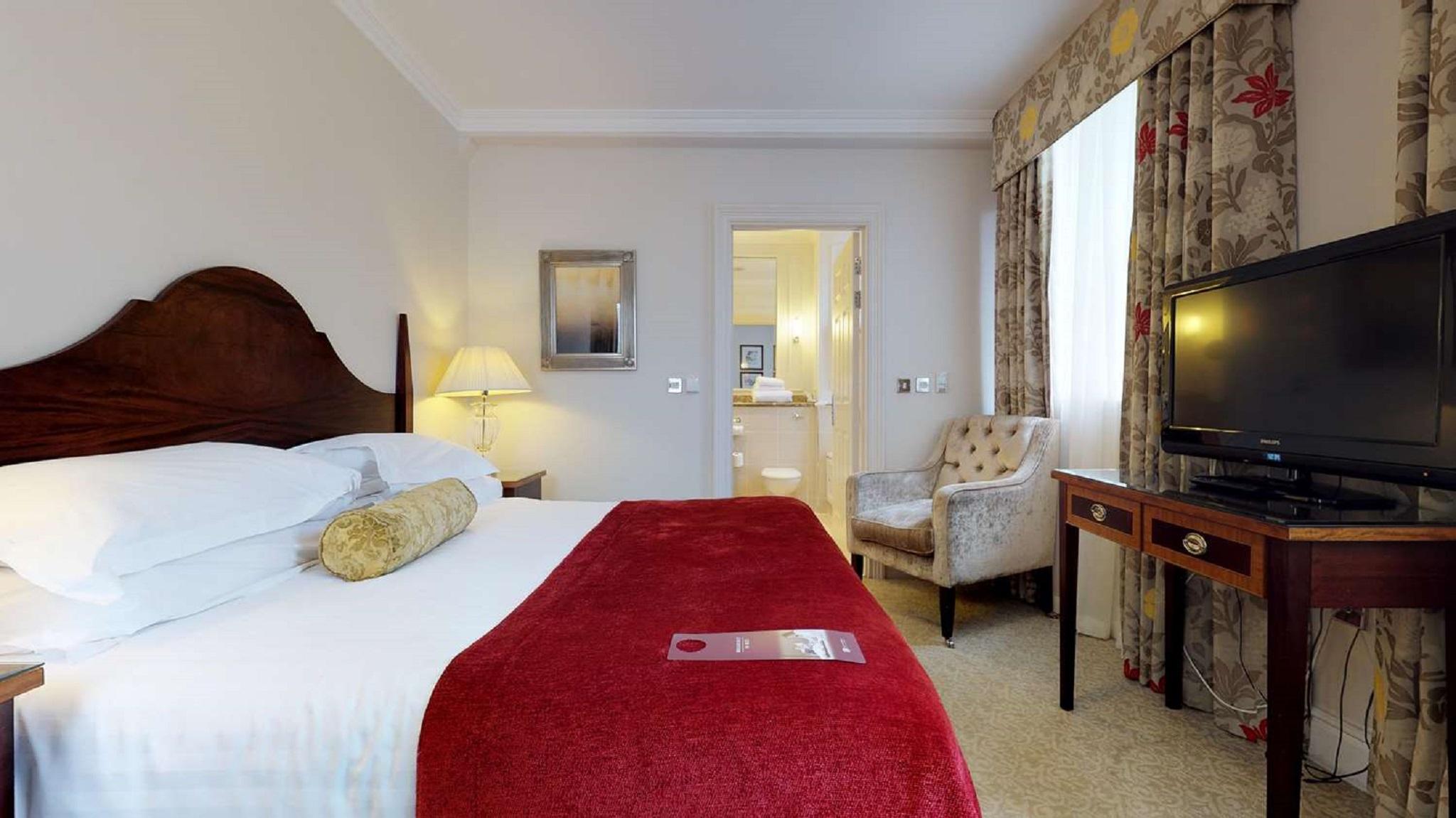 Macdonald Bath Spa Hotel Exterior foto The photo shows a hotel room with a queen-sized bed covered in white linens and a deep red throw blanket. There is a decorative pillow on the bed. Next to the bed, there's a small nightstand with a lamp. In the background, a seating area features a b