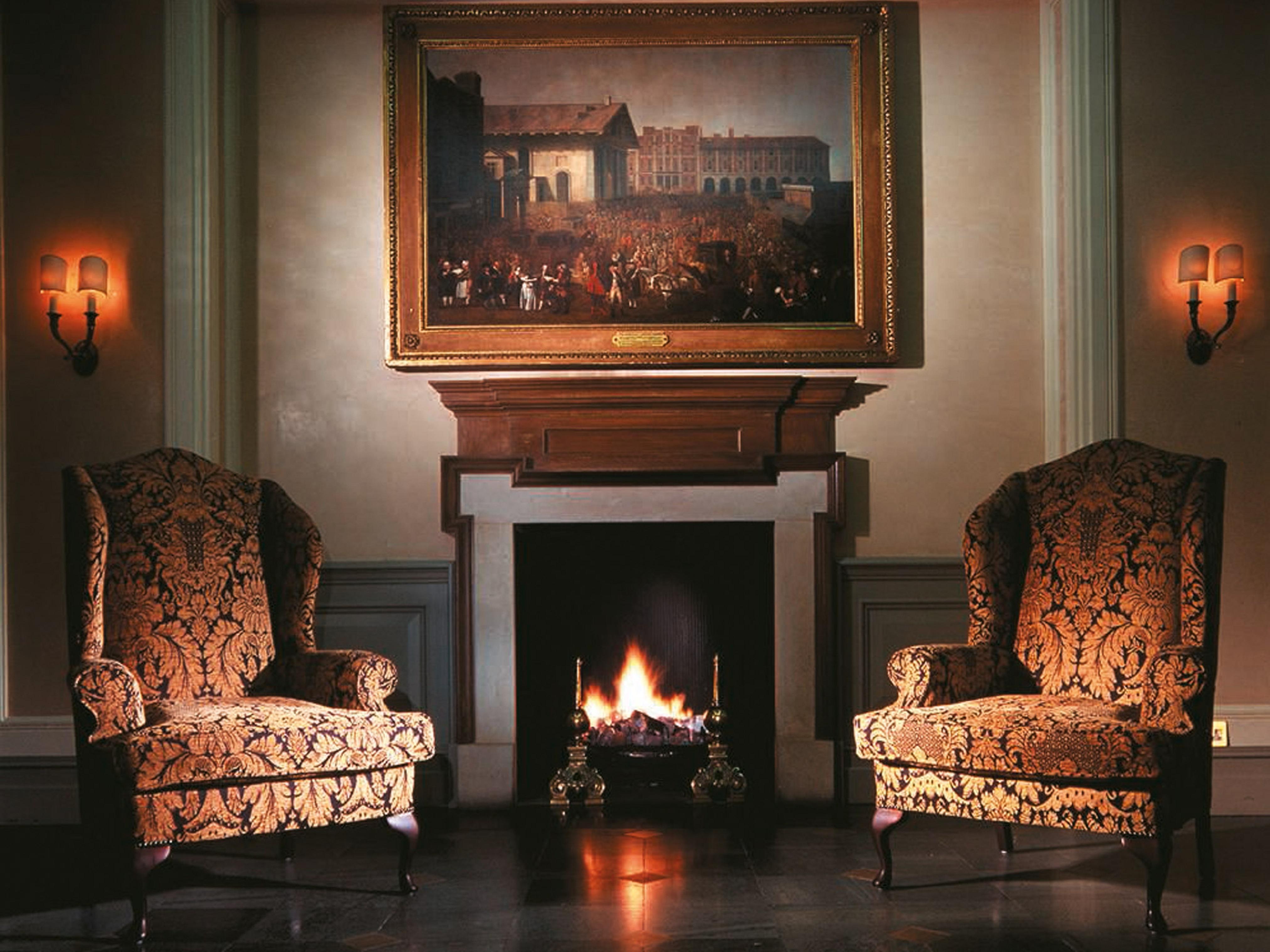 Macdonald Bath Spa Hotel Exterior foto The image shows a cozy indoor setting featuring two ornate armchairs placed opposite a fireplace. Above the fireplace hangs a large painting in a decorative frame. The warm glow from the fire illuminates the room, and there are wall sconces providing