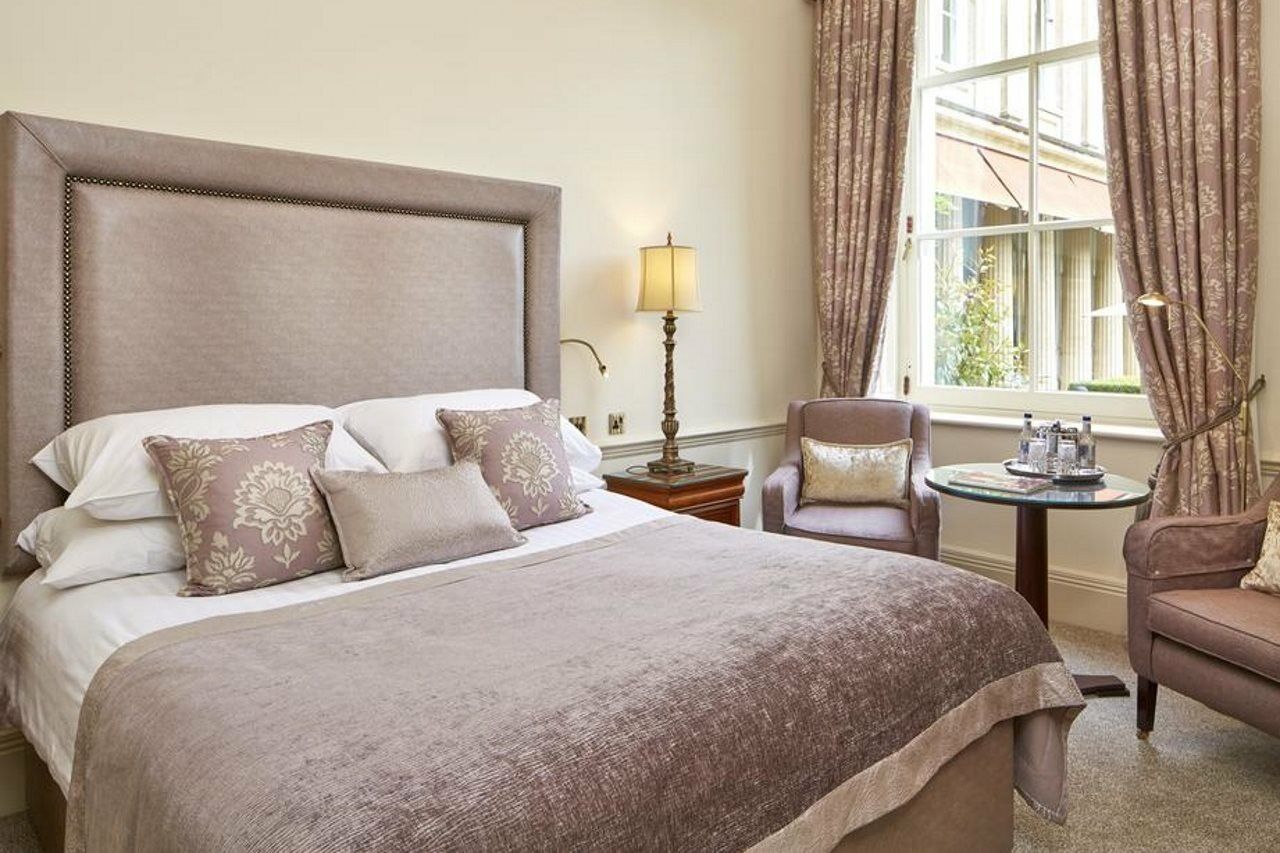 Macdonald Bath Spa Hotel Exterior foto The photo shows a stylish and elegantly decorated hotel room. It features a large bed with soft linens and several decorative pillows. The bedspread is a muted shade of beige or taupe. Beside the bed, there's a small table with two armchairs. One of 